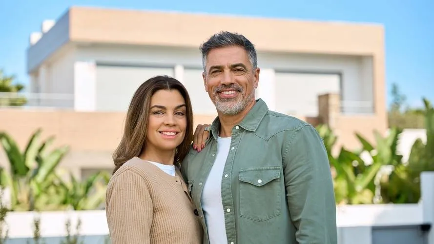 Middle-aged couple smiling, standing outdoors in front of modern beige house with greenery.