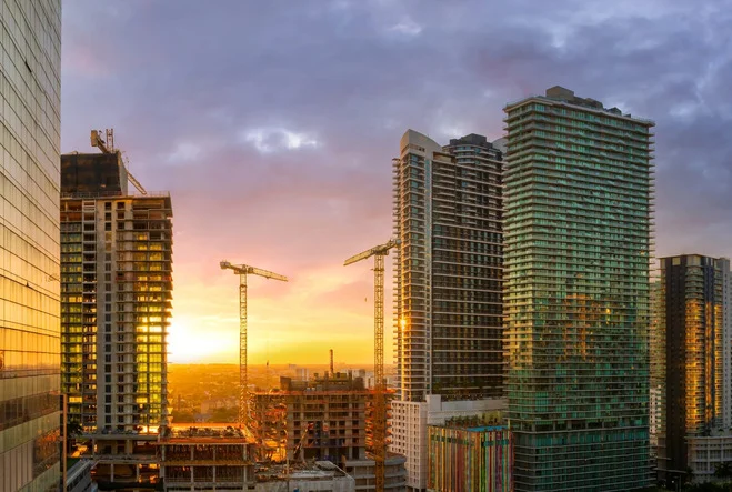Sunset view of a city skyline with high-rise buildings and construction cranes.