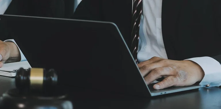 Professional in dark suit typing on laptop with gavel and notepad nearby on desk.