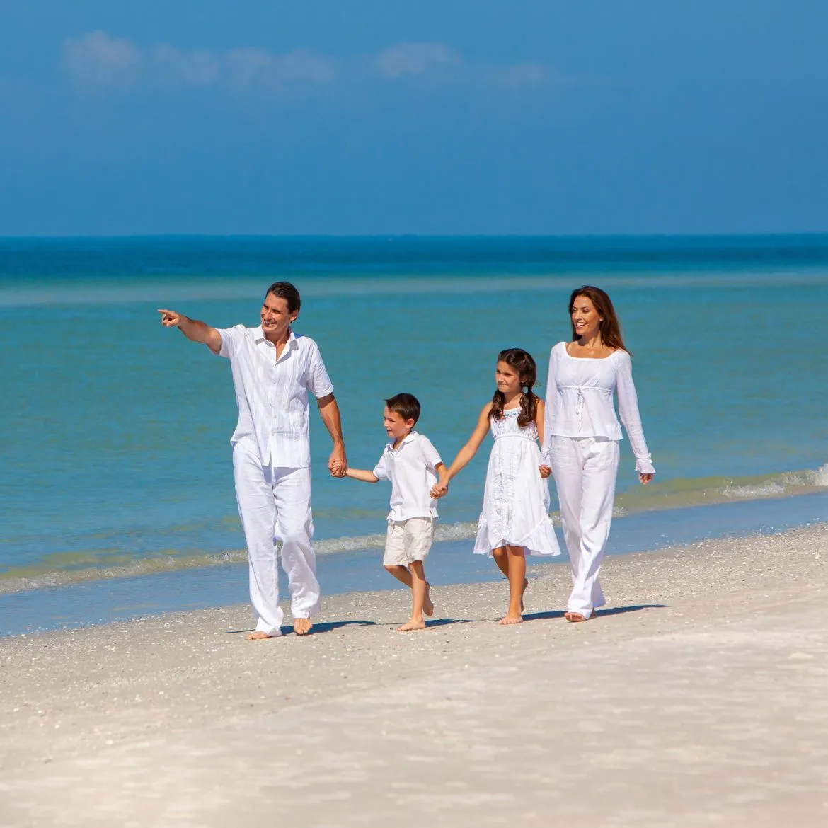 Family of four dressed in white walking barefoot on a sandy beach by the ocean.