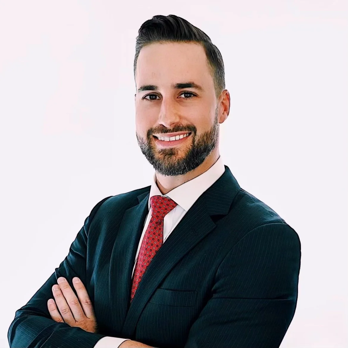 Professional man with short dark hair and beard, wearing a dark suit and red tie, smiling with arms crossed.