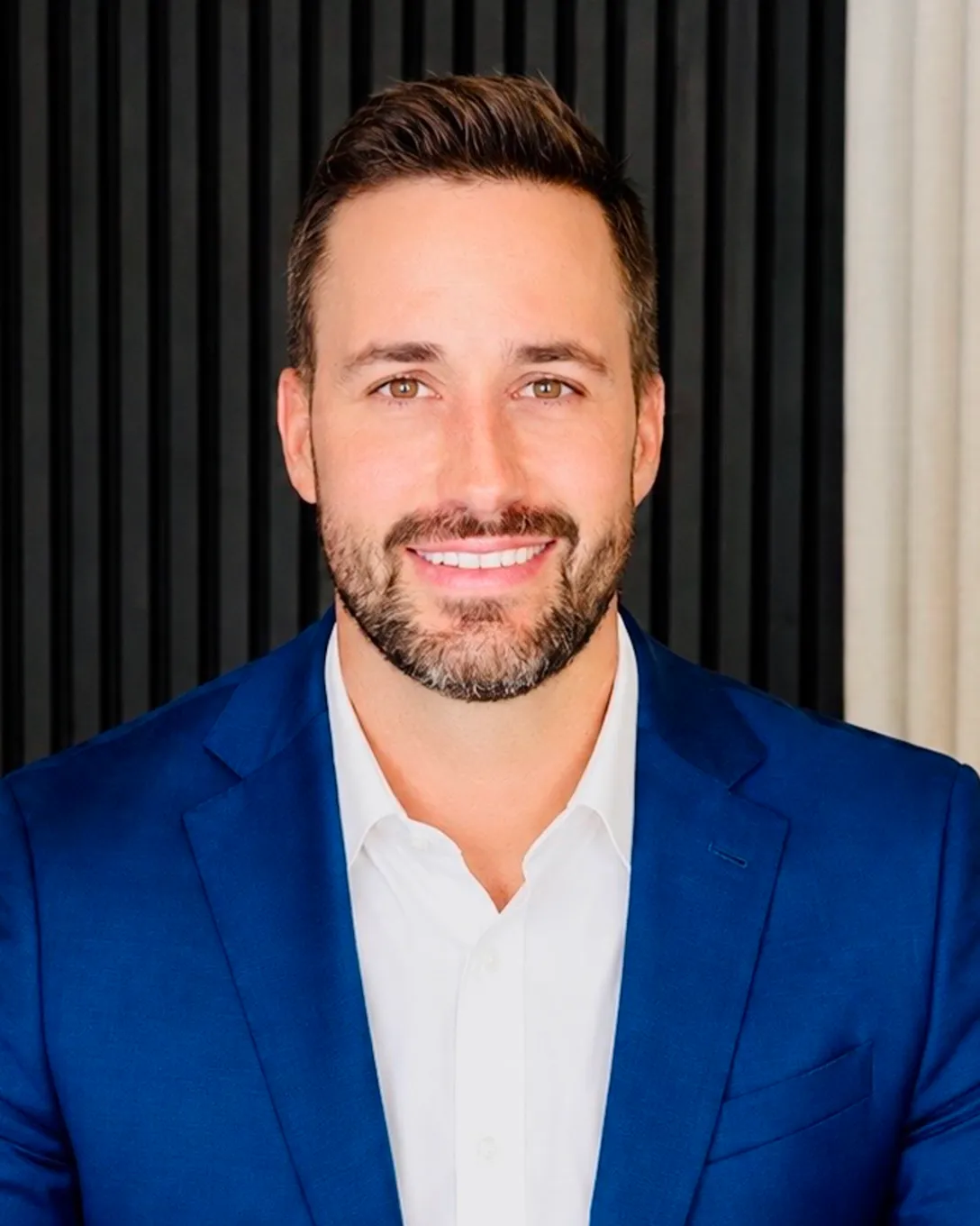 Portrait of a smiling man with brown hair and beard, wearing a blue suit and white shirt.