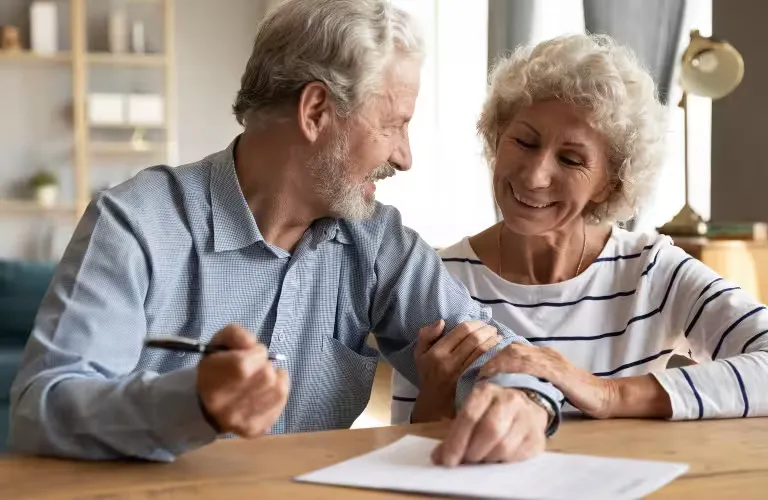 Elderly couple smiling and reviewing documents together at a wooden table.