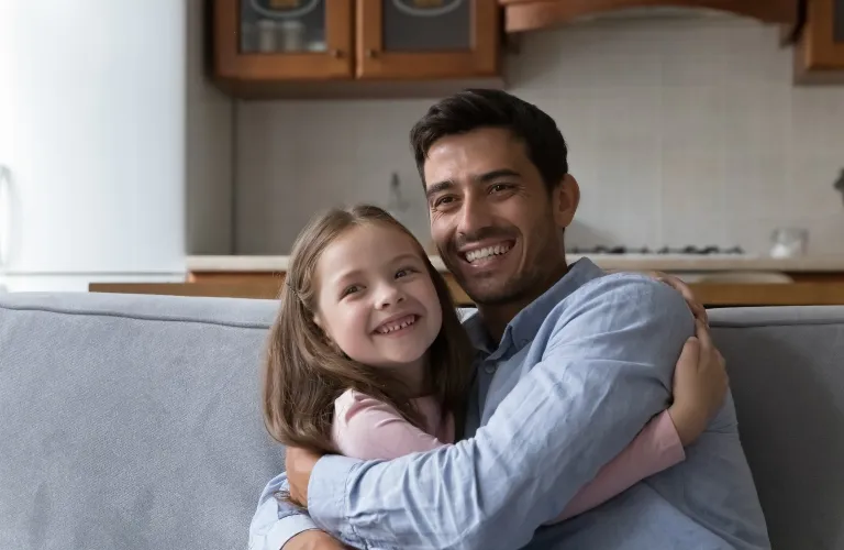 Man and young girl smiling and hugging while seated on a gray couch in a kitchen.