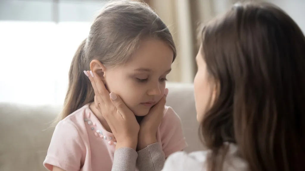 Adult woman gently holding young girl's face, comforting her indoors with soft natural light.